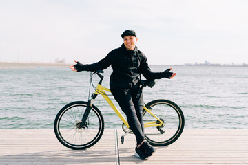 Fototapeta premium a young man in black sportswear stands on a pontoon near the lake and holds a yellow Bicycle near him on a Sunny spring day