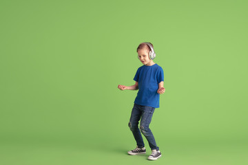 Fototapeta na wymiar Listen to music. Happy boy playing and having fun on green studio background. Caucasian kid in bright looks playful, laughting, smiling. Concept of education, childhood, emotions, facial expression.