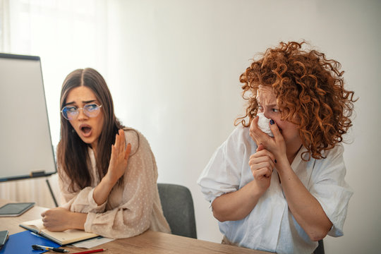 Ill businesswoman blowing and a colleague rejecting her at office. Businesswoman sneezing in tissue near coworker in office. Young businesswoman suffering from a cold and blowing her nose - Powered by Adobe