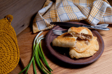 Clay plate of fried meat pies with cutlery and green onion on wooden table.