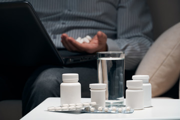 Overworked businessman sitting on the sofa and taking medication. Pills and glass of water near him.