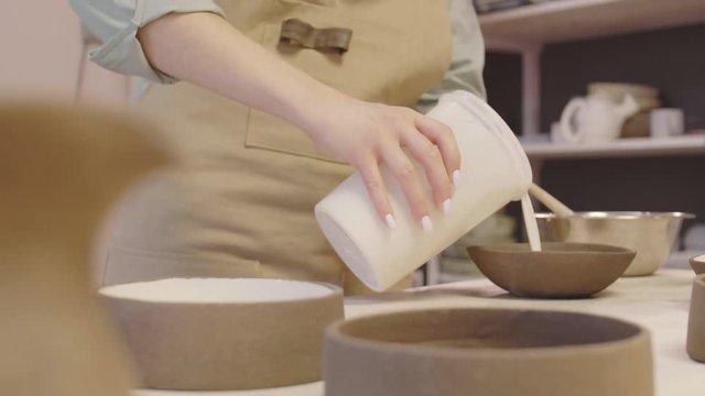 Close up of unrecognizable woman wearing apron pouring white paint from jar into clay plate, spreading paint in plate and then pouring surplus back into jar