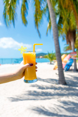 First-person view. Girl holds a glass cup of cold mango fresh on the background of a sandy tropical beach. White sand and palm trees. Fairytale vacation