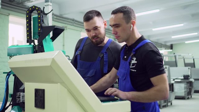 Two male factory workers are discussing near the control panel of a CNC machine. Slider shot.