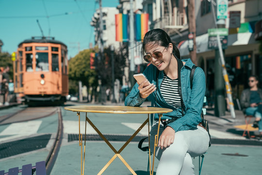 happy beautiful girl tourist sitting at outdoor cafe table and using smart phone chatting online with friends. young woman relax on street with cable car in background. castro district in usa.