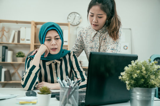 Japanese Girl Colleague Embracing Supporting Islam Woman Reading Bad News In Email On Laptop Computer. Teammate Comforting Stressed Frustrated Female Coworker Upset And Helping To Solve Problem