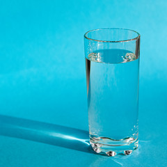 A glass with clean clear clear water stands on a blue background with green shadows. Clean water