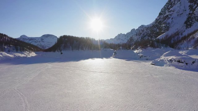 aerial over iced lake covered in snow on mountain valley in sunny winter day.Europe Alps mountains outdoor establisher.4k drone forward flight establishing shot. Sun flare