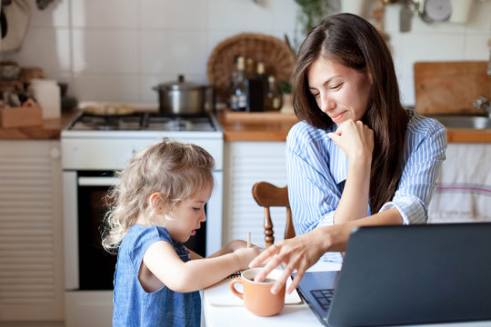 Working mom works from home office with kid. Woman and cute child using laptop. Freelancer workplace in cozy kitchen. Happy mother and daughter. Female business, empathy, care. Lifestyle family moment