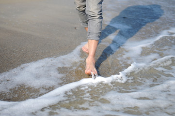 A man is walking along the golden sand of the beach. Male legs walking near the sea. Bare feet of a guy walking along a sandy shore with waves. Summer vacation or vacation.