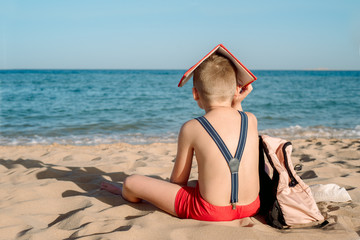 Happy young boy reading a book on a sunny dayl. boy sitting with his back at the beach. Last day of vacation. Back to school. Backpack and suspenders, red book. Bad mood and not a desire to learn.