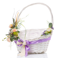 Wicker basket with beautiful decor in green with flowers, greens and wooden branches with purple ribbon around the basket on white background
