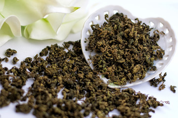 Dry green tea herbs with flowers and vintage porcelain heart shaped plate on white background.