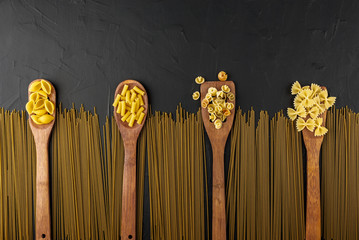 dry pasta on wooden spoons and spaghetti on a textured black background