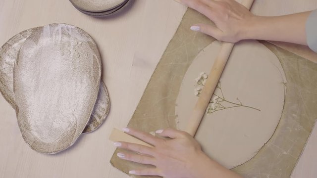 Flat lay of craftswoman hands imprinting beautiful plant on piece of raw clay using wooden rolling pin in ceramic studio