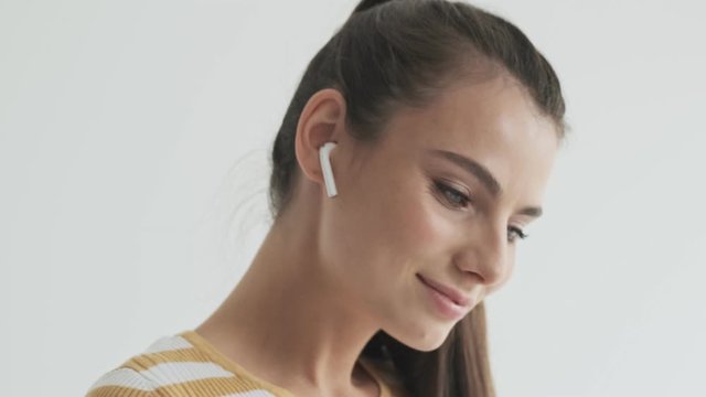A pleased smiling young woman is using her wireless earphones isolated over white background in studio