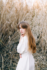 portrait of a young attractive girl in a light white dress and black shoes standing among the reeds in autumn