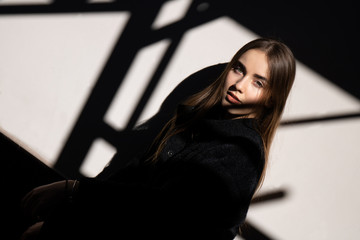 Portrait of young beautiful girl in hard light and shadow with long hair wearing a black coat on white background. Urban. Outdoor. Text space.