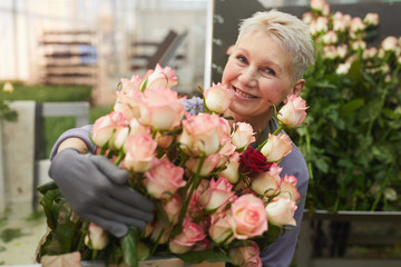 Portrait of happy mature woman with short hair smiling at camera while holding plenty of beautiful roses in her hands