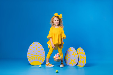 child a blonde girl in a yellow dress is smiling standing with a row of large decorative Easter eggs on a blue background