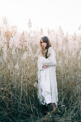 portrait of a young beautiful girl in a white dress, a warm knitted cardigan and black shoes among dry fluffy reeds in the autumn time at sunset