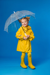 child girl blonde smiling in a yellow raincoat and rubber boots holding an umbrella stands on a blue background in the Studio, space for text