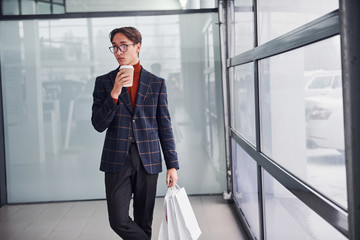 With cup of drink and shopping bags. Young business man in luxury suit and formal clothes is indoors in the office