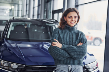 Cheerful young woman standing in front of modern new car indoors