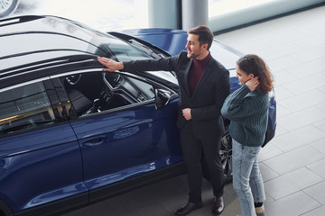 Professional salesman assisting young girl by choosing new modern automobile indoors