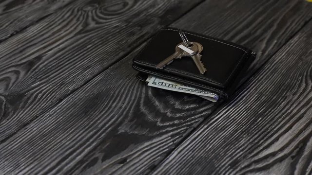 A man puts a wallet on a table of brushed boards. Dollar bills are visible from it. After that puts a bunch of keys on it.