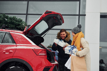 Young couple with gift boxes is near car with tree on the top. Together outdoors at winter time