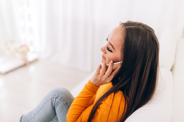 Obraz premium Young girl talking on the phone in an orange sweater on white sofa in white room