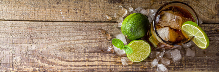 Summer iced alcohol drink with cola and lime. Rum and cola Cuba Libre cocktail. One long glass on wooden background copy space - obrazy, fototapety, plakaty