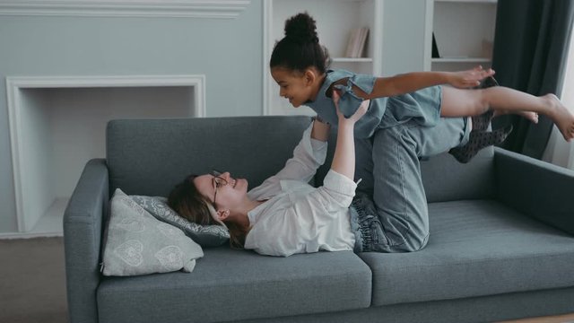 Friendly beautiful family healthy young mother lifting cool child daughter up playing plane on sofa at apartment, funny kid girl having fun together fly in mothers arm practice acro pair yoga bonding