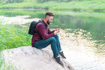 Naklejka na ściany i meble a man with a phone, with a backpack, in a shirt and jeans sits on a stone against the background of nature on a summer day. camping mobile concept