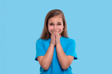 A young girl  crys with grief covering your mouth with palms against blue background in studio