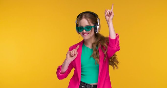 Trendy colorful dressed Woman is dancing in Headphones on yellow Background. Amazing Girl looking happy and satisfied while is listening to Music isolated on Studio. Concept: Musically Apps, Emotions.