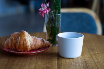 Breakfast in coffee shop. Cup of coffee and fresh croissant in plate. Flowers in glass.