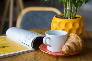 Breakfast in coffee shop. Cup of coffee and fresh croissant in plate. Flowers in glass.