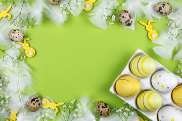 Easter eggs in a white ceramic holder, easter bunny, quail eggs, flowers and white feathers on a green background. Top view. Copy space.