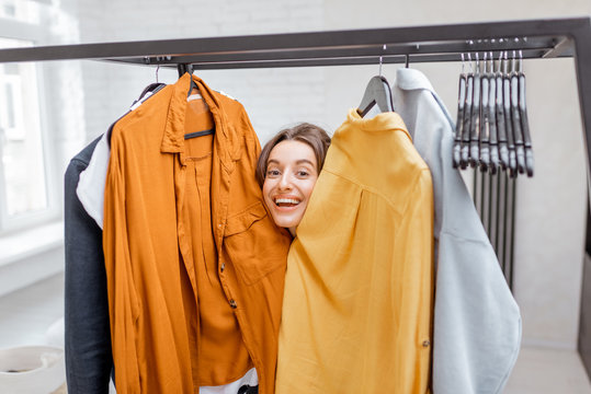 Portrait Of A Young And Cheerful Woman Choosing Casual Clothes To Wear, Standing Between Hangers And Looking Out Of The Clothes In The Wardrobe At The Bedroom