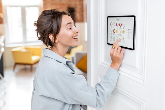 Young woman controlling home with a digital touch screen panel installed on the wall in the living room. Concept of a smart home and mobile application for managing smart devices at home