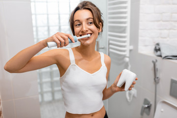 Young and cheerful woman brushing teeth with electric toothbrush during morning hygiene procedures...
