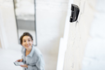 Woman controlling alarm system with a smart phone wireless, standing in the room with motion sensor mounted on the wall