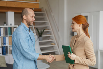Fototapeta premium Young red haired businesswoman in eyeglasses shaking hands with man while they have a meeting at office