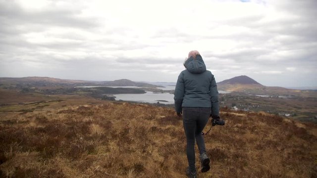 Female photographer holds camera in her hands and walks on grass in Connemara, Ireland, cloudy skies on cold spring day, lake, hills and towns in distance.
