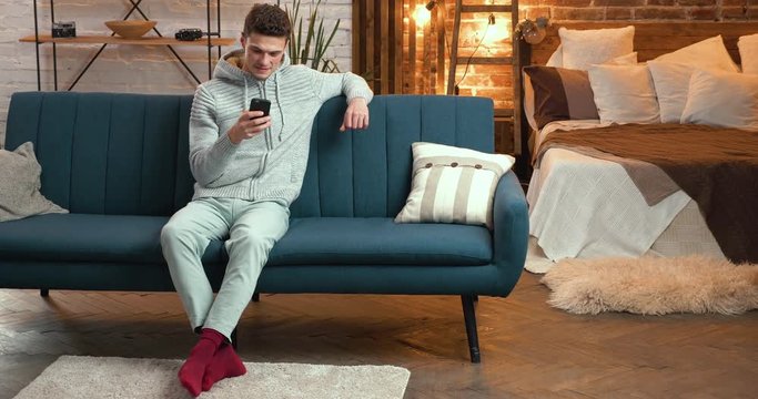 Young Man is sitting in big Loft Flat-Studio and browsing his Smartphone. Looking happy, satisfied. Staring at Home at the Evening, wearing Home Clothes. Apps. Loft Interieur. Using Smartphones.Smile.