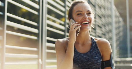 Pretty cheerful Caucasian woman in sporty style talking with smile on smartphone on hot day in morning. Beautiful joyful young girl speaking on mobile phone at sport court. Cellphone talk concept.