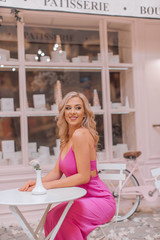 Obraz premium Attractive happy blonde in pink sexy dress sitting at the round cafe table in locations stylized as Paris confectionery