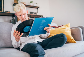 Portrait of young man sitting in comfortable sofa in living room taking out 12-inch vinyl LP from sleeve. Old vintage analog sound value concept image.